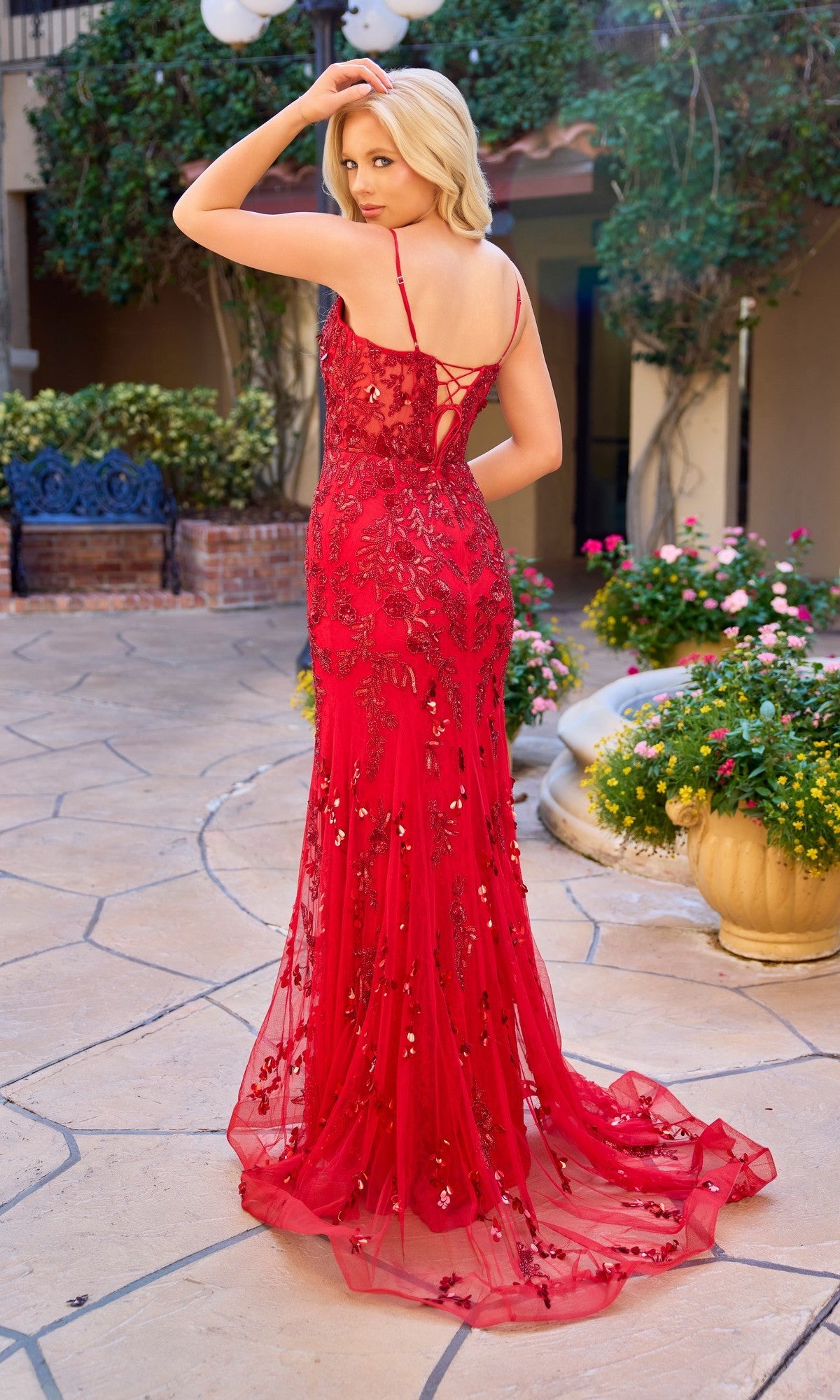 A woman wearing the Primavera 4532 long prom dress stands outdoors on a stone patio, looking over her shoulder and surrounded by flowers and plants.